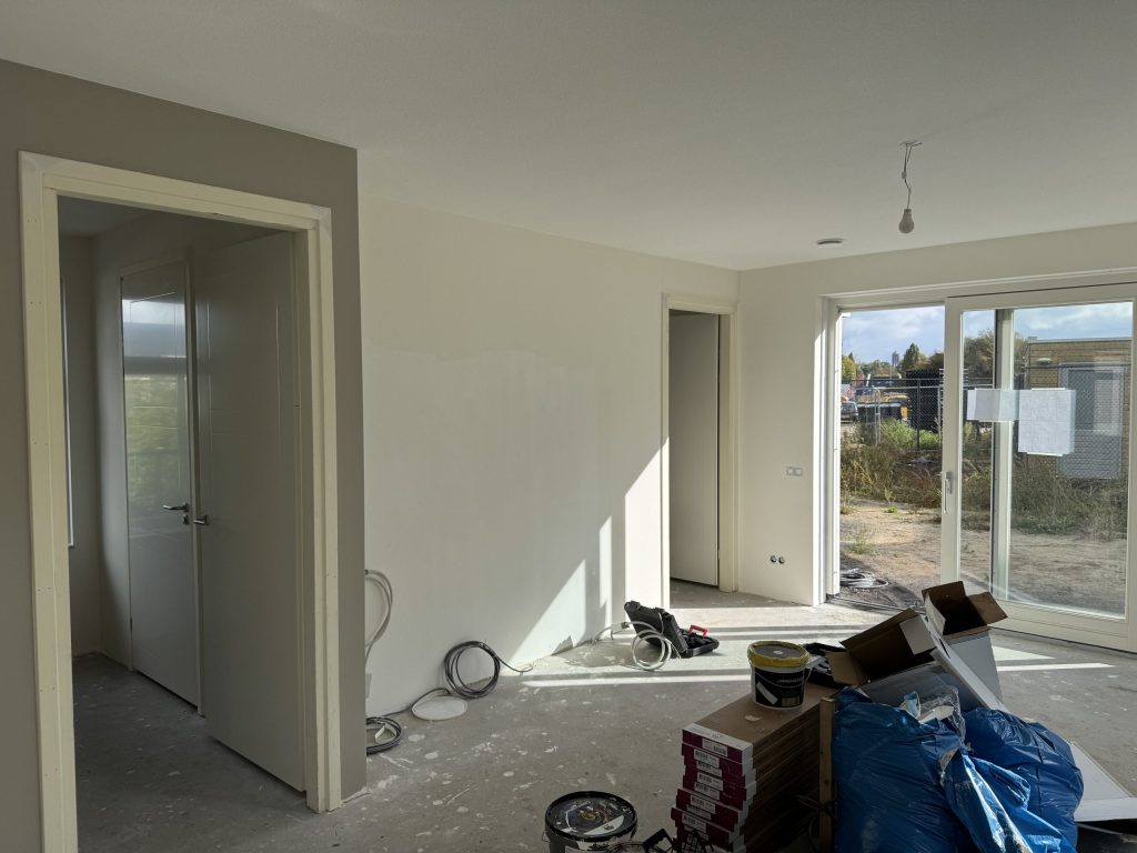 Nieuwbouwwoonkamer tijdens de afwerking, met verfemmers, dozen en kabels op de betonvloer en een tuindeur naar buiten